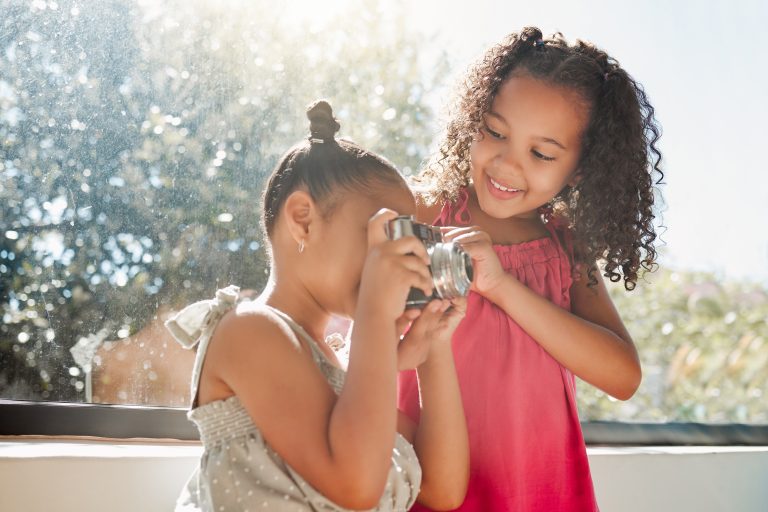 Cute sisters bonding, taking photos on camera at home, smiling while being playful and curious. Lit.